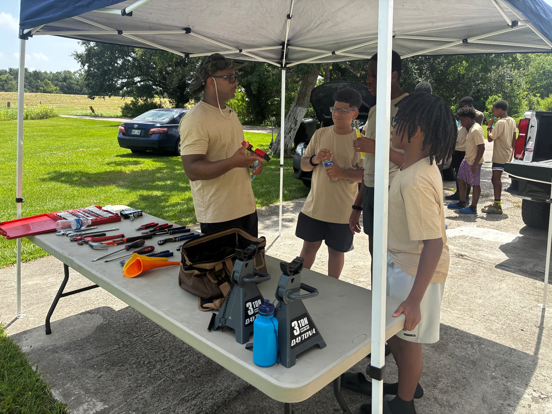A mentor teaching young men basic automotive tools and repair skills under a tent